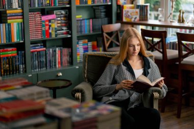 Güzel bir kitapçıda uzun saçlı genç bir kız bir sandalyede oturmuş pasif dinlenme yıllarını okuyor.