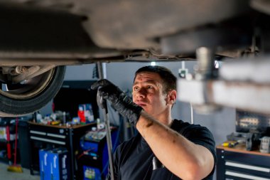 Benzin istasyonundaki oto tamircisi asansörde kaldırılan bir arabayı kontrol eder, mekanik bir araçla arıza düzeltir motor alt gövdenin bir parçasıdır