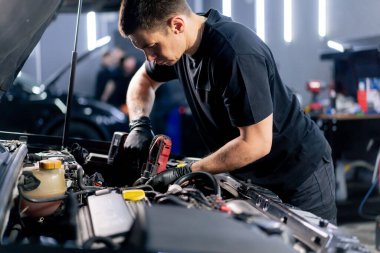 Siyah tişörtlü bir araba servisinde yedek parçaların yerine motor bölümündeki vidaları sökmek için matkap kullanır.