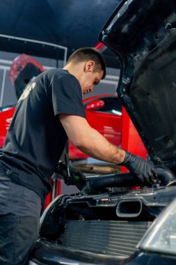 Bir araba hizmet merkezinde siyah tişörtlü bir usta araç gereçleri kullanır ve yedek parçaları değiştirerek motor bölümündeki vidaları söker.