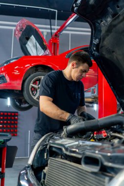 Bir araba hizmet merkezinde siyah tişörtlü bir usta araç gereçleri kullanır ve yedek parçaları değiştirerek motor bölümündeki vidaları söker.