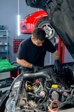 Bir araba hizmet merkezinde siyah tişörtlü bir usta araç gereçleri kullanır ve yedek parçaları değiştirerek motor bölümündeki vidaları söker.