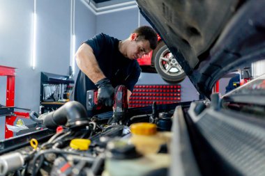 Siyah tişörtlü bir araba servisinde yedek parçaların yerine motor bölümündeki vidaları sökmek için matkap kullanır.