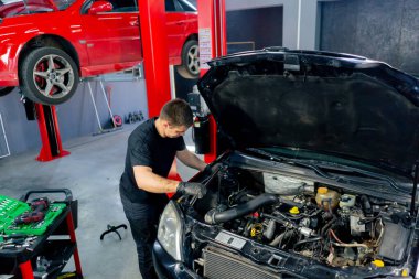 Bir araba hizmet merkezinde siyah tişörtlü bir usta araç gereçleri kullanır ve yedek parçaları değiştirerek motor bölümündeki vidaları söker.