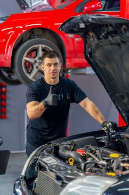 Bir araba servisinde siyah tişörtlü ve eldivenli bir usta açık kapüşonlu bir arabanın yanında duruyor kameraya bakıyor ve kırmızı bir arabanın arka planına hafifçe gülümsüyor.