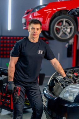 Bir araba servisinde siyah tişörtlü ve eldivenli bir usta açık kapüşonlu bir arabanın yanında duruyor kameraya bakıyor ve kırmızı bir arabanın arka planına hafifçe gülümsüyor.