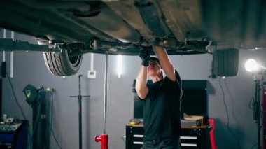 Benzin istasyonundaki oto tamircisi asansörde kaldırılan bir arabayı kontrol eder, mekanik bir araçla arıza düzeltir motor alt gövdenin bir parçasıdır