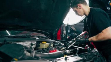 Siyah tişörtlü bir araba servisinde yedek parçaların yerine motor bölümündeki vidaları sökmek için matkap kullanır.