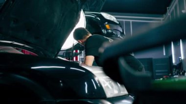 Siyah tişörtlü bir araba servisinde yedek parçaların yerine motor bölümündeki vidaları sökmek için matkap kullanır.