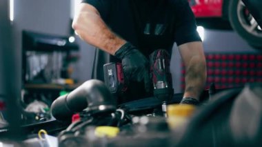 Siyah tişörtlü bir araba servisinin kapağını kapatın. Yedek parçaları değiştirmek için motor bölümündeki vidaları sökmek için matkap kullanın.