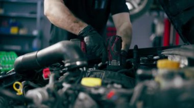 Siyah tişörtlü bir araba servisinde yedek parçaların yerine motor bölümündeki vidaları sökmek için matkap kullanır.