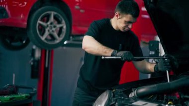 Siyah tişörtlü bir araba servisinde yedek parçaların yerine motor bölümündeki vidaları sökmek için matkap kullanır.