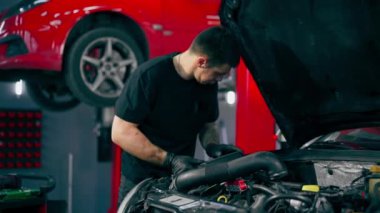 Siyah tişörtlü bir araba servisinde yedek parçaların yerine motor bölümündeki vidaları sökmek için matkap kullanır.