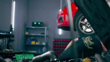 Siyah tişörtlü bir araba servisinde yedek parçaların yerine motor bölümündeki vidaları sökmek için matkap kullanır.