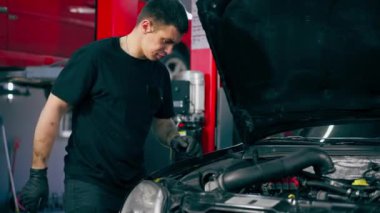 Siyah tişörtlü bir araba servisinde yedek parçaların yerine motor bölümündeki vidaları sökmek için matkap kullanır.