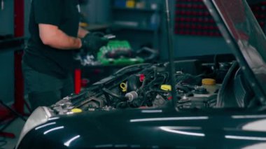Siyah tişörtlü bir araba servisinde yedek parçaların yerine motor bölümündeki vidaları sökmek için matkap kullanır.