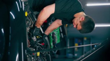 Siyah tişörtlü bir araba servisi ustasındaki dikey video yedek parçaların yerine arabanın motor bölümündeki vidaları sökmek için matkap kullanır.
