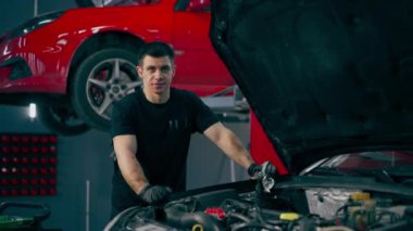 Bir araba servisinde siyah tişörtlü ve eldivenli bir usta açık kapüşonlu bir arabanın yanında duruyor kameraya bakıyor ve kırmızı bir arabanın arka planına hafifçe gülümsüyor.