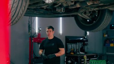 Benzin istasyonundaki oto tamircisi asansörde kaldırılan bir arabayı kontrol eder, mekanik bir araçla arıza düzeltir motor alt gövdenin bir parçasıdır