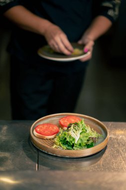 Profesyonel bir mutfakta, temiz metal bir masanın üzerinde kadın bir şef taze etle salata toplar ve yuvarlak ahşap bir tabakta mozzarella ile tost yapar.