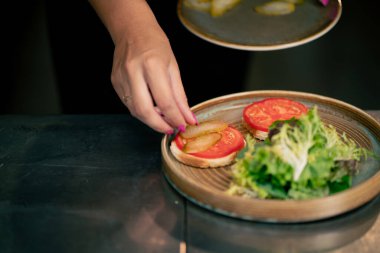 Profesyonel bir mutfakta, temiz metal bir masanın üzerinde kadın bir şef taze etle salata toplar ve yuvarlak ahşap bir tabakta mozzarella ile tost yapar.