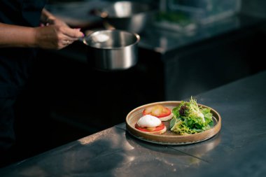 Profesyonel bir mutfakta, temiz metal bir masanın üzerinde kadın bir şef taze etle salata toplar ve yuvarlak ahşap bir tabakta mozzarella ile tost yapar.