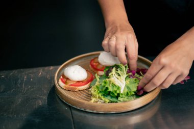 Profesyonel bir mutfakta, temiz metal bir masanın üzerinde kadın bir şef taze etle salata toplar ve yuvarlak ahşap bir tabakta mozzarella ile tost yapar.