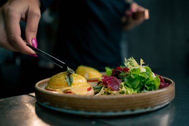 Profesyonel bir mutfakta, temiz metal bir masanın üstünde kadın bir şef taze etle bir salata toplar ve ahşap bir tabakta mozzarella ile tost yapar.