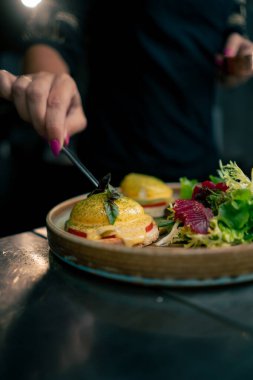 Profesyonel bir mutfakta, temiz metal bir masanın üstünde kadın bir şef taze etle bir salata toplar ve ahşap bir tabakta mozzarella ile tost yapar.