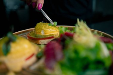 Profesyonel bir mutfakta, temiz metal bir masanın üstünde kadın bir şef taze etle bir salata toplar ve ahşap bir tabakta mozzarella ile tost yapar.