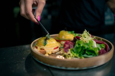 Profesyonel bir mutfakta, temiz metal bir masanın üstünde kadın bir şef taze etle bir salata toplar ve ahşap bir tabakta mozzarella ile tost yapar.