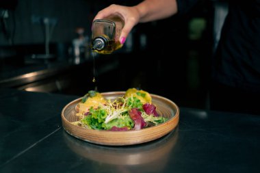 Profesyonel bir mutfakta, temiz metal bir masanın üstünde kadın bir şef taze etle bir salata toplar ve ahşap bir tabakta mozzarella ile tost yapar.