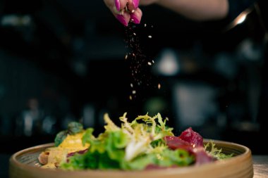 Profesyonel bir mutfakta, temiz metal bir masanın üstünde kadın bir şef taze etle bir salata toplar ve ahşap bir tabakta mozzarella ile tost yapar.