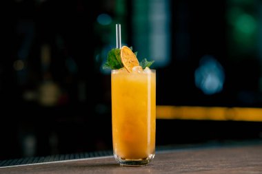 Koyu bir arka planda yakın plan. Nane ve kurutulmuş portakalla süslenmiş sarı-turuncu karışımlı uzun cam bir bardak.