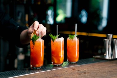 Koyu bir arka planda üç uzun cam bardağa nane ve kurutulmuş turuncu barmenle süslenmiş sarı-turuncu bir kokteyl kokteyle pipet yerleştirir.