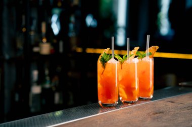 Koyu bir arka planda üç uzun cam bardakta, nane ve kurutulmuş portakalla süslenmiş sarı-turuncu bir kokteyl.