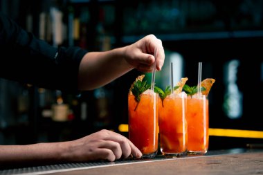 Koyu bir arka planda üç uzun cam bardağa nane ve kurutulmuş turuncu barmenle süslenmiş sarı-turuncu bir kokteyl kokteyle pipet yerleştirir.