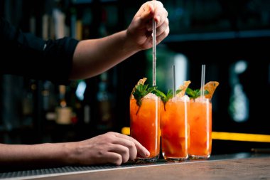 Koyu bir arka planda üç uzun cam bardağa nane ve kurutulmuş turuncu barmenle süslenmiş sarı-turuncu bir kokteyl kokteyle pipet yerleştirir.