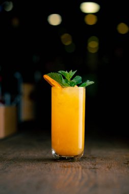 Koyu bir arka planda yakın plan. Nane ve kurutulmuş portakalla süslenmiş sarı-turuncu karışımlı uzun cam bir bardak.