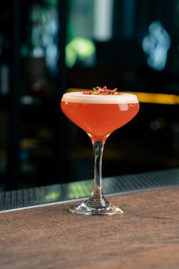 Karanlık arka planda bir barda cam bir bardak var. Cam üzerine köpüklü kırmızı bir kokteyl damlıyor. Portakal dilimi ve kırmızı baharat.