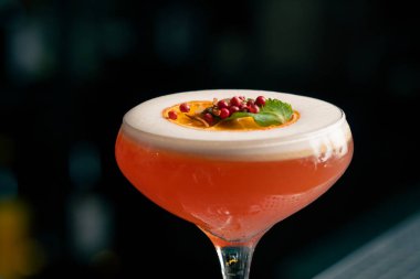 Koyu bulanık arka plandaki bir barda cam bir cam bardak var. Camda köpüklü kırmızı bir kokteyl damlaları var. Bir portakal dilimi ve kırmızı baharat.