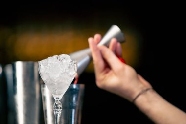 Erkek barmen içeceği ustalıkla çelik jigger 'ın içine dolduruyor. Yanında da ezilmiş buzla birlikte uzun şeffaf bir bardak var.