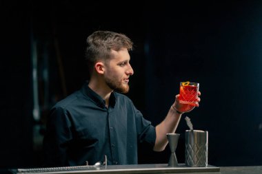 Siyah arkaplanda bir barda siyah gömlekli yakışıklı bir barmen hazır kırmızı bir erkek kokteyli ile tezgahın üzerinde duruyor.