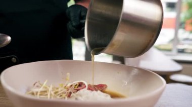 Fast food restoranındaki dikey videoda siyah üniformalı büyük bir şef Japon udon çorbasını hazır malzemelerden hazırlar ve çorba döker.