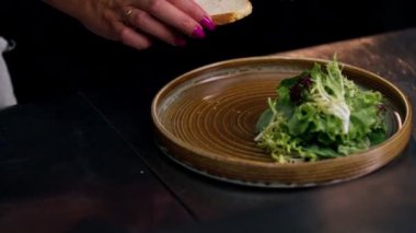 Profesyonel bir mutfakta, temiz metal bir masanın üzerinde kadın bir şef taze etle bir salata toplar ve yuvarlak ahşap bir tabakta mozzarella ile tost yapar.