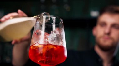 Siyah bir zemin üzerinde şarap kadehini kapatıyoruz. Camda ezilmiş buz var. Portakal rengi Aperol Spritz bir bardağa dökülüyor.
