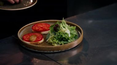 Profesyonel bir mutfakta, temiz metal bir masanın üzerinde kadın bir şef taze etle bir salata toplar ve yuvarlak ahşap bir tabakta mozzarella ile tost yapar.