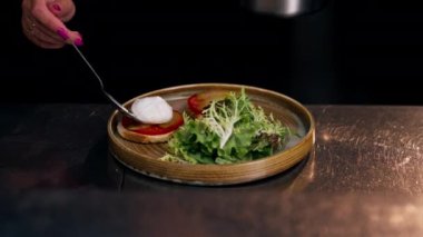 Profesyonel bir mutfakta, temiz metal bir masanın üzerinde kadın bir şef taze etle bir salata toplar ve yuvarlak ahşap bir tabakta mozzarella ile tost yapar.