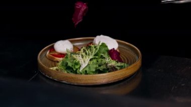 Profesyonel bir mutfakta, temiz metal bir masanın üzerinde kadın bir şef taze etle bir salata toplar ve yuvarlak ahşap bir tabakta mozzarella ile tost yapar.
