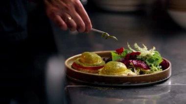 Profesyonel bir mutfakta, temiz metal bir masanın üzerinde kadın bir şef taze etle bir salata toplar ve yuvarlak ahşap bir tabakta mozzarella ile tost yapar.